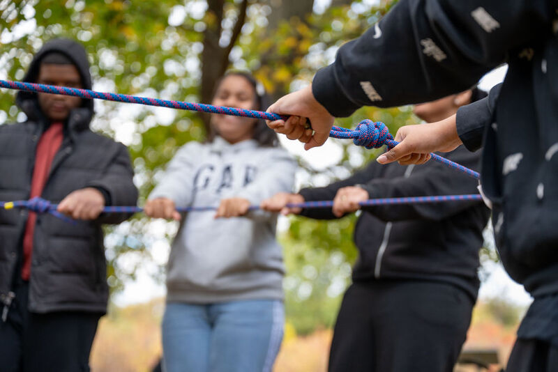 The image shows a group of people holding a rope, likely participating in a team-building activity. They appear to be outdoors, possibly in a park or wooded area, with trees visible in the background. The focus is on the hands and the rope, suggesting collaboration and coordination. The people seem engaged and focused on the task at hand.